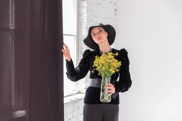 Junges Mädchen weibliches Modell Brünette mit schwarzem Hut mit einem Strauß gelber Blumen - Foto, Bild