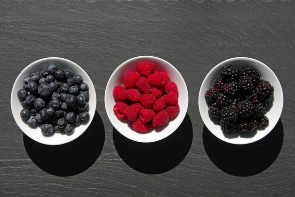 Top view of three white porcelain bowls on dark slate with fresh blueberries, raspberries, and blackberries - Fotó, kép