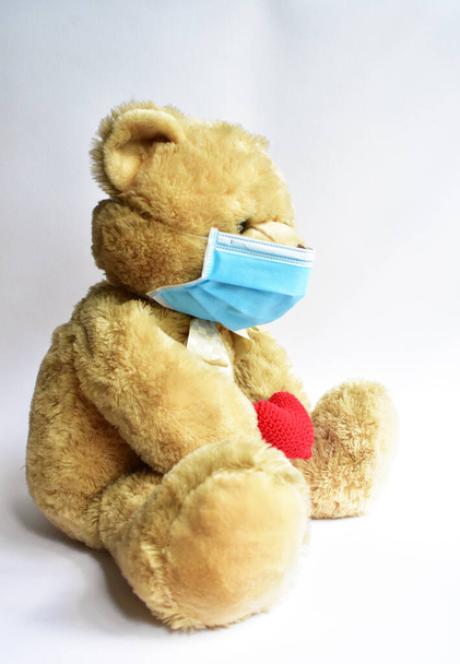 Söpö ruskea karhu sininen lääketieteellinen naamio valkoisella taustalla, punainen sydän. Teddy karhu yllään kasvonaamio suojaava leviämisen taudin virus CoV-2 Corona virus tauti karanteeni, pysyä turvassa - Valokuva, kuva
