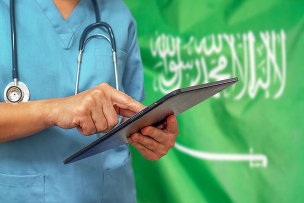 Chirurg of arts die een digitale tablet gebruikt op de achtergrond van de Saoedi-Arabische vlag. Medische apparatuur of medisch netwerk, technologie en diagnostiek in Saoedi-Arabië. - Foto, afbeelding
