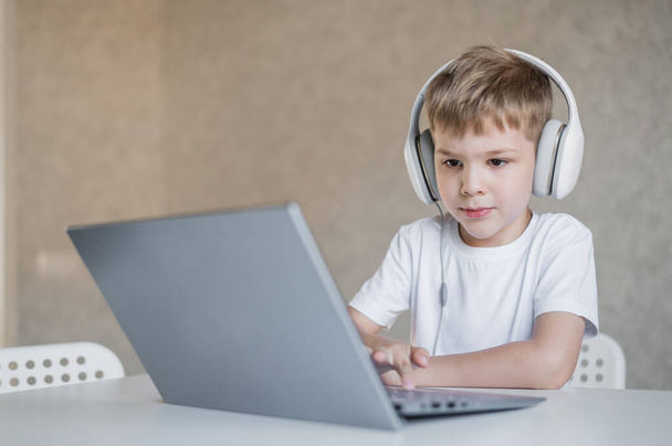 Cute little boy is watching cartoons or a movie on a laptop. Caucasian child sits at a table at home and listens to music on headphones. - 写真・画像