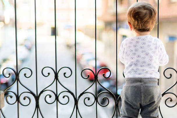 Un niño de un año y medio jugando en el balcón y viendo pasar los coches en cuarentena se aburre porque no puede salir.
 - Foto, imagen