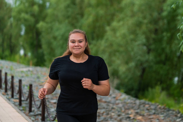 An obese young woman jogging outdoors. Fat beautiful smiling girl in a black tracksuit is engaged in fitness for weight loss on the waterfront. A woman runs on a summer day. - Foto, Imagem