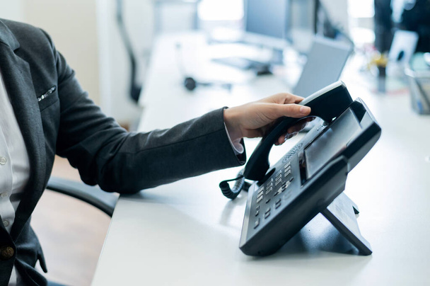 Closeup female hand on landline phone in office. Faceless woman in a suit works as a receptionist answering the phone to customer calls. - Φωτογραφία, εικόνα