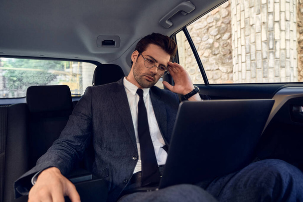 Jovem empresário confiante seriamente e atenciosamente olhando para o laptop segurando óculos com uma mão enquanto sentado no carro
. - Foto, Imagem