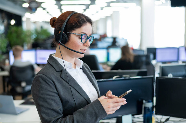 Beautiful caucasian woman in headset is holding a mobile phone while standing in open space office. Friendly female helpdesk operator browsing the screen of a smartphone in the workplace. - Фото, изображение