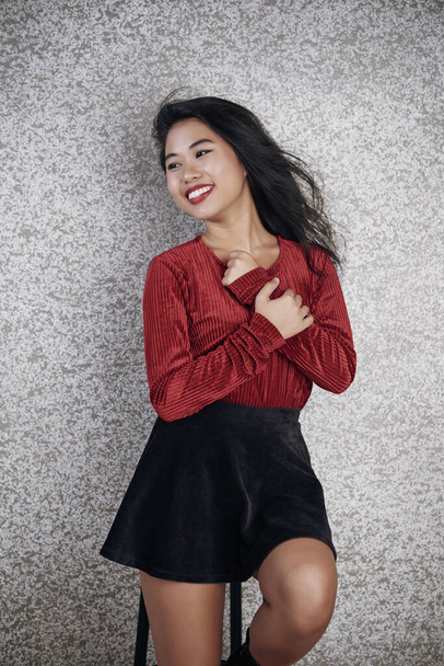 Encantadora mujer asiática vestida con elegante atuendo sentada en el taburete de la barra mirando hacia otro lado y sonriendo, fondo gris de la pared
 - Foto, imagen
