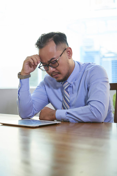 Потрясающий азиатский мужчина в очках, сидящий за рабочим столом и читающий важные документы с помощью цифрового планшета - Фото, изображение