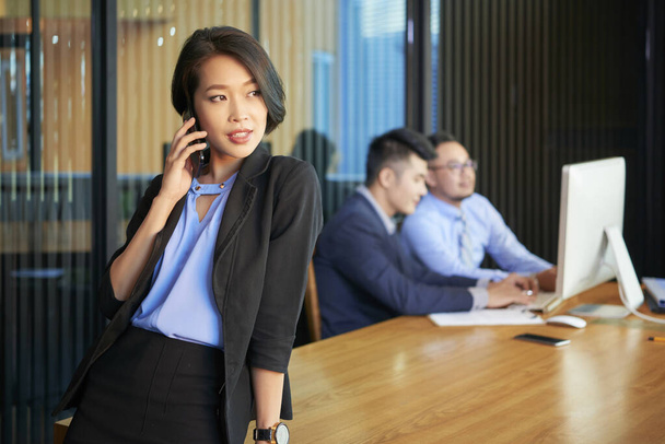Bonita mulher de negócios asiática em pé à mesa no escritório moderno falando ao telefone olhando para longe, tiro horizontal
 - Foto, Imagem