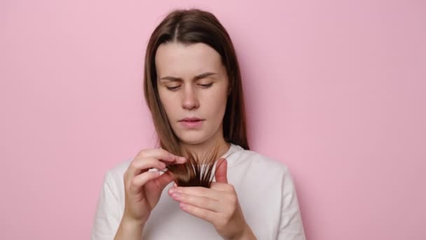 Depressed young woman looking at split ends, isolated on pink studio background. Female hormone problems or vitamin deficiency. Unhappy girl feels upset about brittle damaged dry hair loss concept - Πλάνα, βίντεο