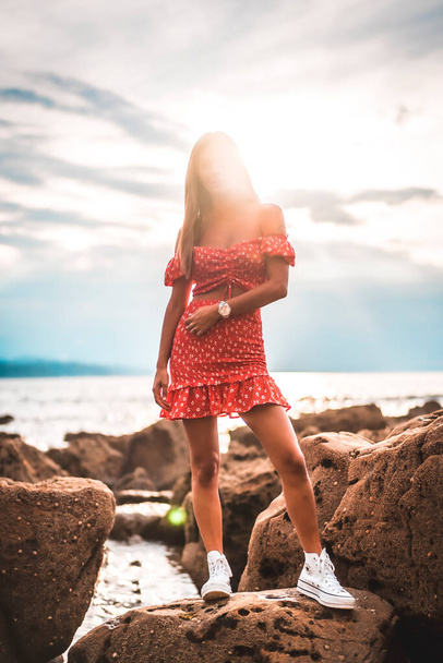 A young brunette Caucasian woman in a red dress on the beach of Itzurrun in the town of Zumaia, Gipuzkoa. Basque Country. Lifestyle session, on top of a standing rock with the sun in the background - Photo, image