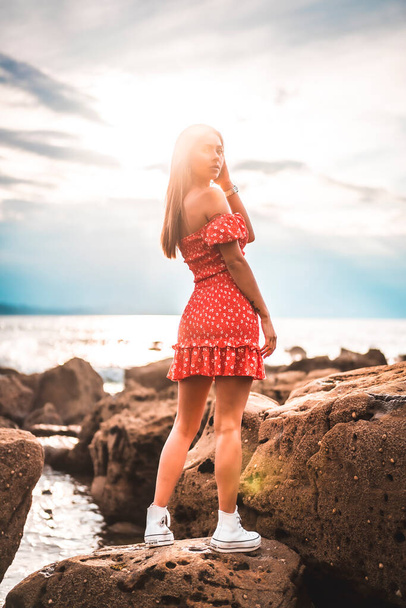 A young brunette Caucasian woman in a red dress on the beach of Itzurrun in the town of Zumaia, Gipuzkoa. Basque Country. Lifestyle session, on top of a rock with the sun in the background - Zdjęcie, obraz