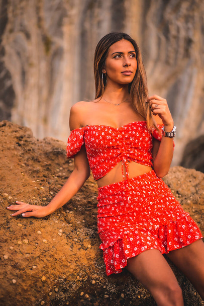 A young brunette Caucasian woman in a red dress on the beach of Itzurrun in the town of Zumaia, Gipuzkoa. Basque Country. Lifestyle session, portrait sitting on a green sea rock looking to the right - Photo, Image