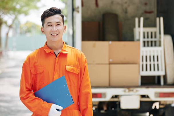 Portrait horizontal moyen de jeune homme asiatique en mouvement portant un uniforme orange vif debout à l'extérieur tenant un presse-papiers bleu, espace de copie - Photo, image