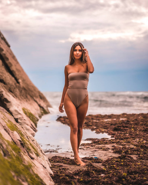 A dark-haired caucasian woman in a brown swimsuit on a natural background by the sea in the town of Zumaia, Gipuzkoa. Basque Country. In the summer sunset. Pretty young woman walking on the on coast - Foto, imagen