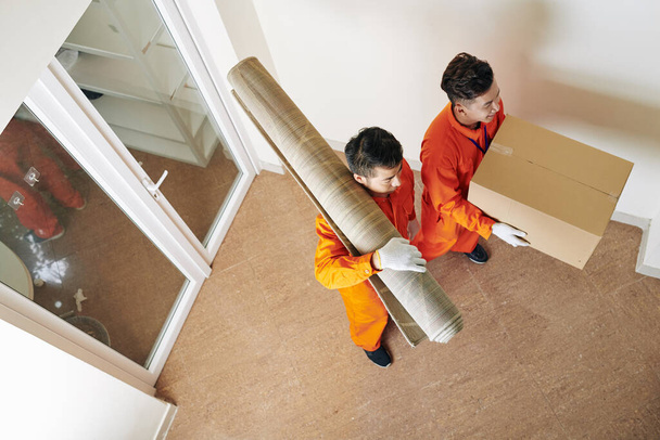 High angle view shot of two moving men wearing orange uniform carrying carpet and box with household things - Foto, Imagen
