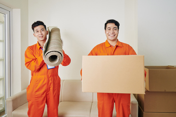 Веселые азиатские мужчины в оранжевой форме, держащие ковер и коробку с бытовыми вещами, смотрящие в камеру, горизонтальный средний выстрел - Фото, изображение