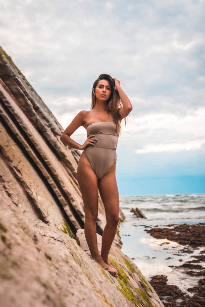 A dark-haired caucasian woman in a brown swimsuit on a natural background by the sea in the town of Zumaia, Gipuzkoa. Basque Country. In the summer sunset. On top of some rocks with open arms - Fotografie, Obrázek