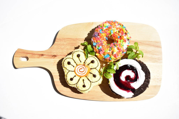 Три барвисті пончики в дерев'яній обробній дошці, сонячно-білий фон, листя м'яти, нездорове харчування, фастфуд, смачні, смачні, солодкі, солодкі частування, зручна їжа, приготування їжі, домашнє
 - Фото, зображення