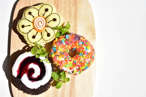 Drie kleurrijke donuts in houten snijplank, zonnige witte achtergrond, muntblaadjes, junkfood, fastfood, lekker, heerlijk, zoet, suikerzoet, handig eten, koken, zelfgemaakt - Foto, afbeelding