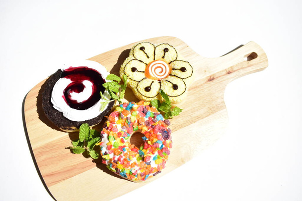 Три красочных пончика в деревянной доске резки, солнечный белый фон, мятные листья, фаст-фуд, вкусные, вкусные, сладкие, сладкие лакомства, удобная еда, приготовление пищи, домашнее - Фото, изображение