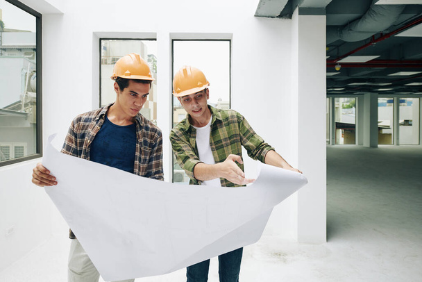 Two professional engineers wearing checked shirts and hardhats working on construction plan, horizontal medium long shot - Photo, Image