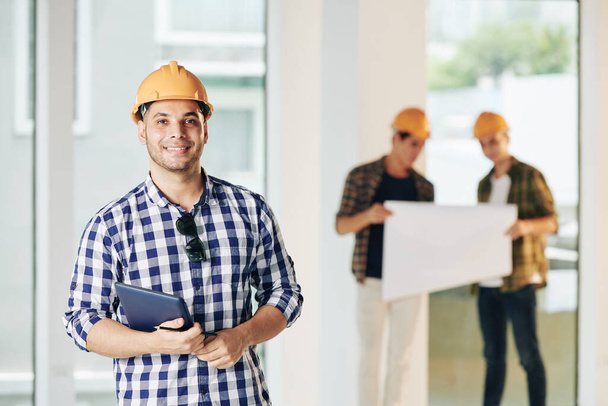 Medium portrait of professional engineer wearing casual shirt and hardhat holding tablet looking at camera, his colleagues looking at building plan - Photo, Image