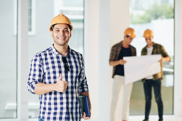 Medium portrait of construction engineer wearing casual shirt and hardhat looking at camera showing thumb up, his colleagues looking at building plan - Photo, Image