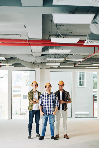 Vertical tiro longo de três engenheiros profissionais vestindo camisas verificadas e hardhats de pé juntos dentro de novo espaço de construção
 - Foto, Imagem