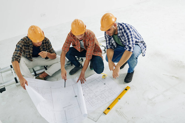 Horizontal high angle shot of modern construction workers sitting on hunkers looking into building plan papers, copy space - Photo, Image