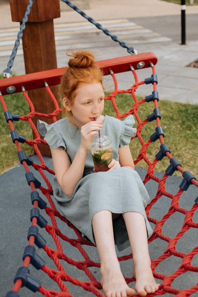 Rudowłosa dziewczyna huśtająca się w hamaku ze szklanką zimnej lemoniady. - Zdjęcie, obraz