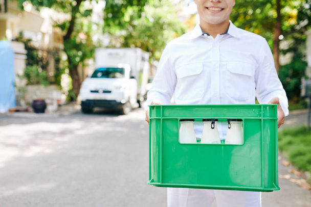Horizontal head cropped shot of delivery man wearing white uniform clothes standing outdoors holding box with milk bottles, copy space - Photo, Image