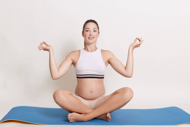 Young beautiful pregnant yoga model working out indoors, expectant mother sitting in yogic cross-legged pose against white wall, looks smiling at camera. Prenatal meditation. - Zdjęcie, obraz