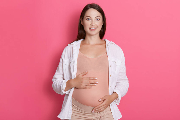 Pregnant female wearing casual attire looking at camera with charming smile, keeping hands on belly, being happy, expectant mother with dark hair expressing positive emotions. - Foto, afbeelding
