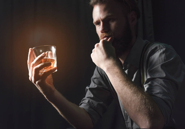 Гарний молодий чоловік з бородою сидить у темній кімнаті зі склянкою алкоголю в руках. Поняття відпочинку і релаксації, бузку і самотності
 - Фото, зображення