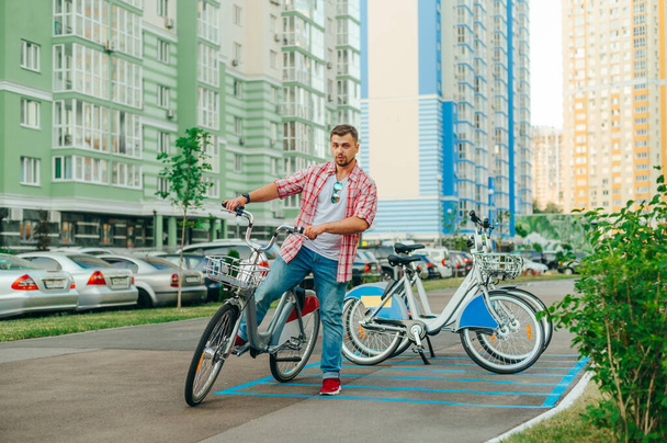 Συμπυκνωμένος άντρας στέκεται με ποδήλατα στο φόντο μιας σειράς ποδηλάτων προς ενοικίαση και ποζάρει για την κάμερα. Ένας άνθρωπος περπατά στην πόλη με οικολογικές μεταφορές. - Φωτογραφία, εικόνα