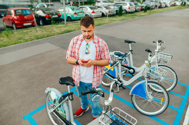 明るいシャツの髭の男は、モバイルアプリケーションを介して自転車を借りて、路上の自転車の行の近くに深刻な顔にスマートフォンを使用しています - 写真・画像