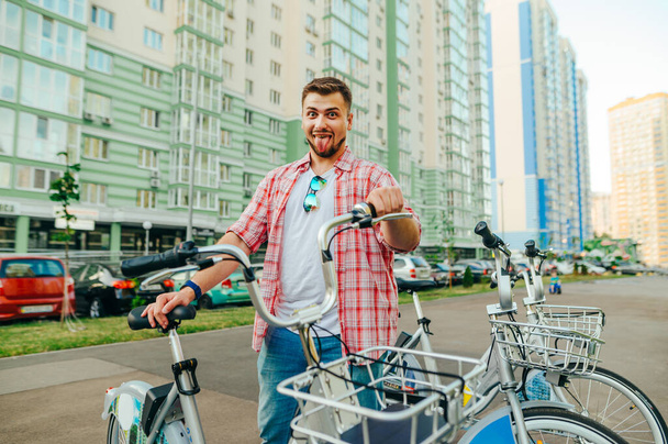 Funny young man stands with a rented bicycle against the backdrop of the cityscape and makes a funny face, looks at the camera and smiles. Cheerful man takes a sharing bike from a row. - Photo, Image