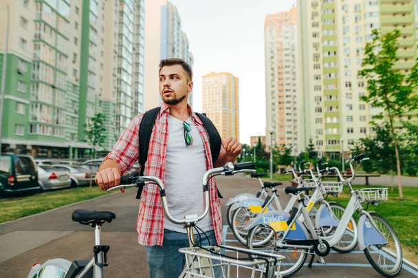 Όμορφος ενήλικας με πουκάμισο και σακίδιο στέκεται με φόντο το αστικό τοπίο και μια σειρά από ποδήλατα, κοιτάζοντας αλλού με σοβαρό πρόσωπο. Ένας άντρας με νοικιασμένο ποδήλατο περπατάει. - Φωτογραφία, εικόνα