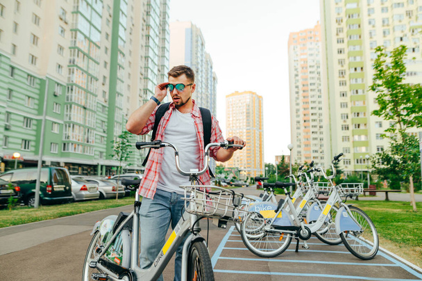 A cool adult man walks around the city on a rented bicycle, wears sunglasses and looks away against the backdrop of the cityscape. The tourist picked up a bicycle and posed for the camera - Photo, Image