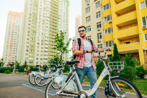 Ένας χαρούμενος νεαρός περπατάει στο δρόμο με ένα νοικιασμένο ποδήλατο πόλης, κοιτάζει αλλού και χαμογελάει. Ο άνθρωπος χρησιμοποιεί τις οικολογικές μεταφορές σε μια απογευματινή βόλτα. - Φωτογραφία, εικόνα