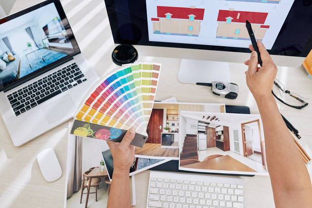 Σχεδιαστής εσωτερικών χώρων που εργάζεται στο σχέδιο χρώματος για το εσωτερικό και εξωτερικό του σπιτιού πελατών, κρατώντας την παλέτα χρωμάτων και δείχνοντας στην οθόνη lap-top με stylus - Φωτογραφία, εικόνα