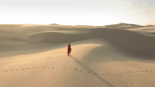 4K εναέρια άποψη ενός κοριτσιού σε φτερουγίσματα κόκκινο φόρεμα περπάτημα σε αμμόλοφους στο ηλιοβασίλεμα - Πλάνα, βίντεο