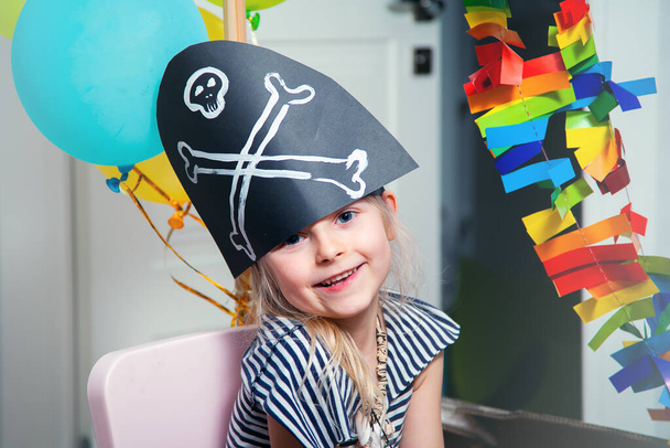 A cheerful little child in a pirate costume plays at home on a cardboard sea ship with a black flag. Fun games at home with family. Travels and adventures at home - Photo, Image
