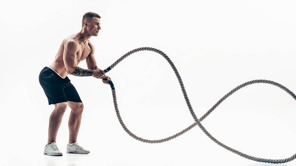 Attractive muscular man working out with heavy ropes. Photo of handsome man in sportswear isolated on white background - Foto, afbeelding