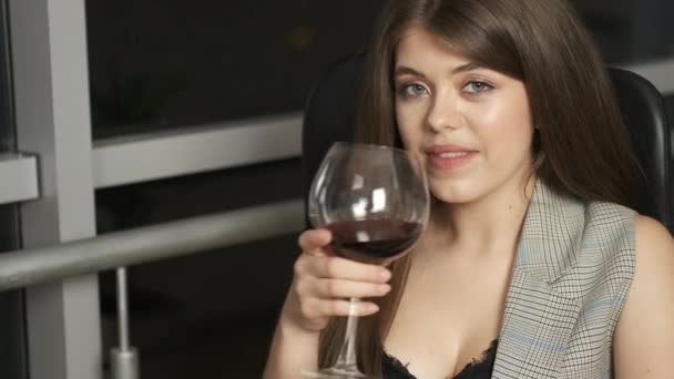Beautiful girl of Caucasian appearance with long hair sitting by the panoramic window reads a book holding a glass of red wine and enjoys the night city - Video