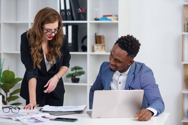 Two diverse businesspeople chatting sitting behind laptop in office. Excited caucasian female sharing ideas or startup business plan with black male coworker. Informal conversation, work break concept - 写真・画像