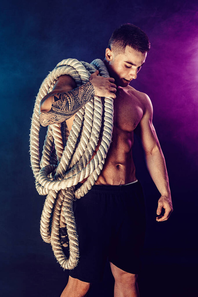 Bearded athletic looking bodybulder holding battle rope on dark studio background with smoke. Strength and motivation - Photo, Image