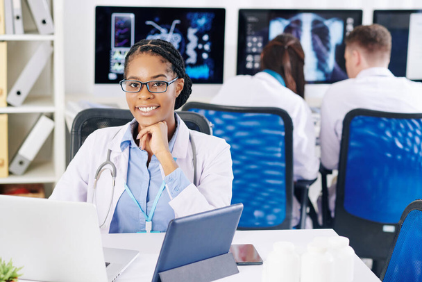 Retrato de jovem médico sorridente em óculos sentado na mesa de escritório e trabalhando em laptop
 - Foto, Imagem