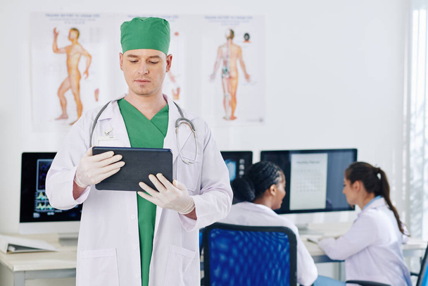 Súlyos sebész olvassa kórtörténetét beteg táblagépen, kollégái megvitatása munkarend a háttérben - Fotó, kép